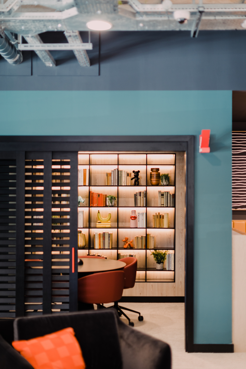 A modern interior with a bookshelf filled with books and decor items, partially visible through a sliding wooden door. The room has teal walls, brown leather chairs, and chic, industrial elements like exposed ductwork.
